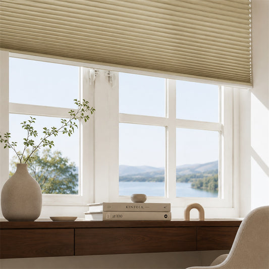Beige pleated window blinds installed on a white-framed window, shown in a bright room with a view of a lake and mountains. A wooden desk with books, a vase with branches, and part of a chair are visible below.