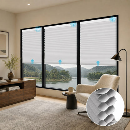 A set of white cellular roller blinds installed on a large black-framed window in a modern living room. The image includes blue arrow overlays indicating airflow and a circular inset showing a close-up of the textured honeycomb pattern of the blinds.