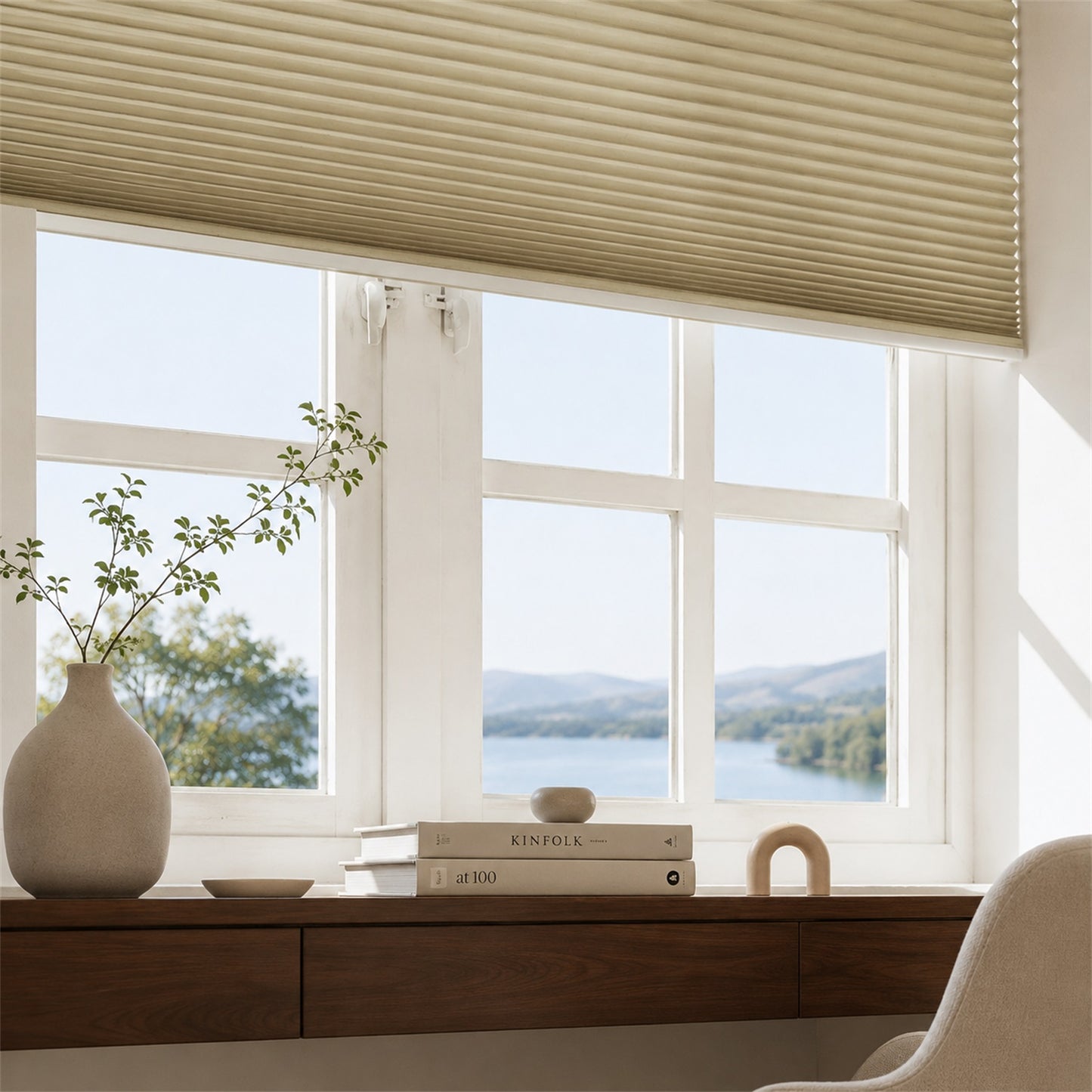 Beige pleated window blinds installed on a white-framed window, shown in a bright room with a view of a lake and mountains. A wooden desk with books, a vase with branches, and part of a chair are visible below.