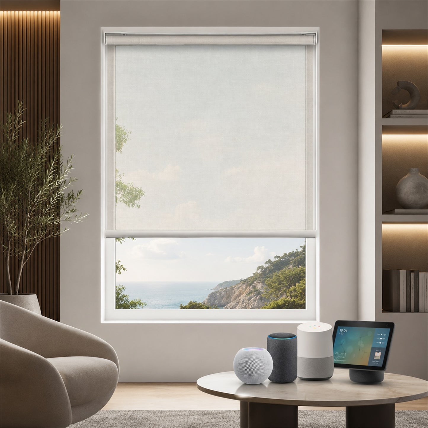 A modern roller blind in a light beige or off-white shade, partially drawn on a large window overlooking a scenic coastal landscape. The room setting includes a beige armchair, a round wooden coffee table with smart home devices, and built-in wooden shelving with warm lighting.