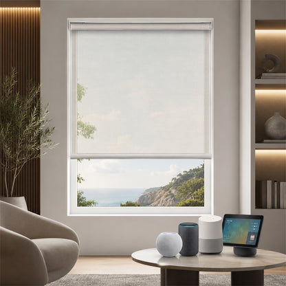 A modern roller blind in a light beige or off-white shade, partially drawn on a large window overlooking a scenic coastal landscape. The room setting includes a beige armchair, a round wooden coffee table with smart home devices, and built-in wooden shelving with warm lighting.