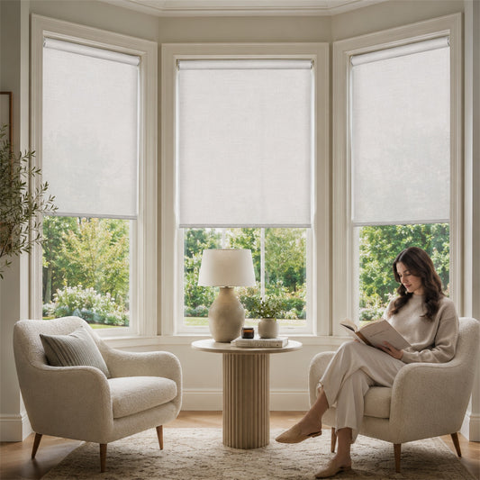 A woman sits in a cream upholstered armchair in a bright corner room, reading a book beside a small round table with a lamp and potted plants. Two more matching armchairs are visible, and the windows above have light-colored roller shades partially drawn.