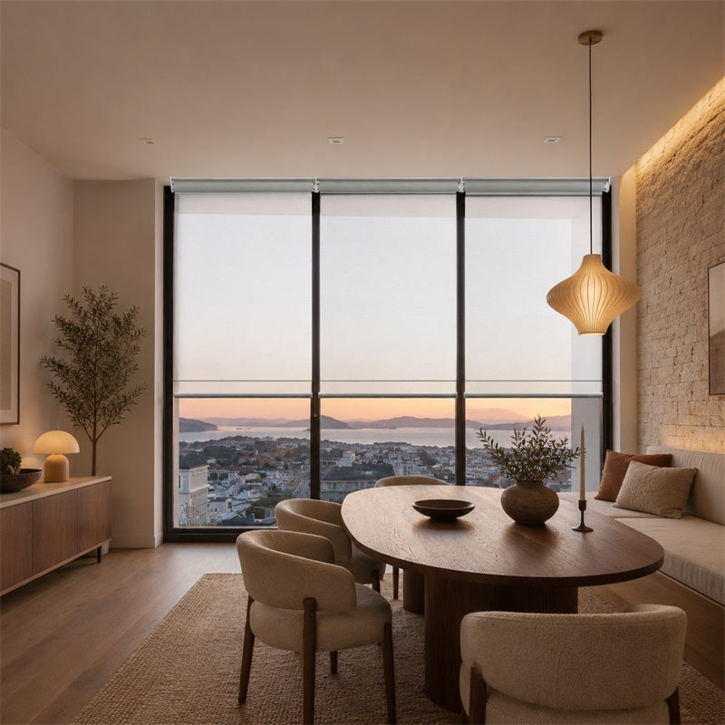 A modern dining room interior with a round wooden dining table, upholstered chairs, a pendant light, a built-in sofa, and large floor-to-ceiling windows overlooking a coastal city at sunset.